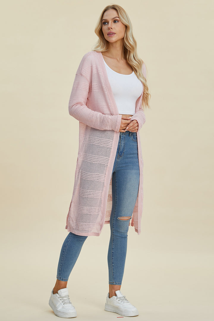 A woman with long blonde hair is elegantly dressed in a white top and the Double Take Full Size Open Front Longline Cardigan in light pink. She pairs it with ripped blue jeans made from stretchy fabric, and her look is completed with white sneakers against the beige background.