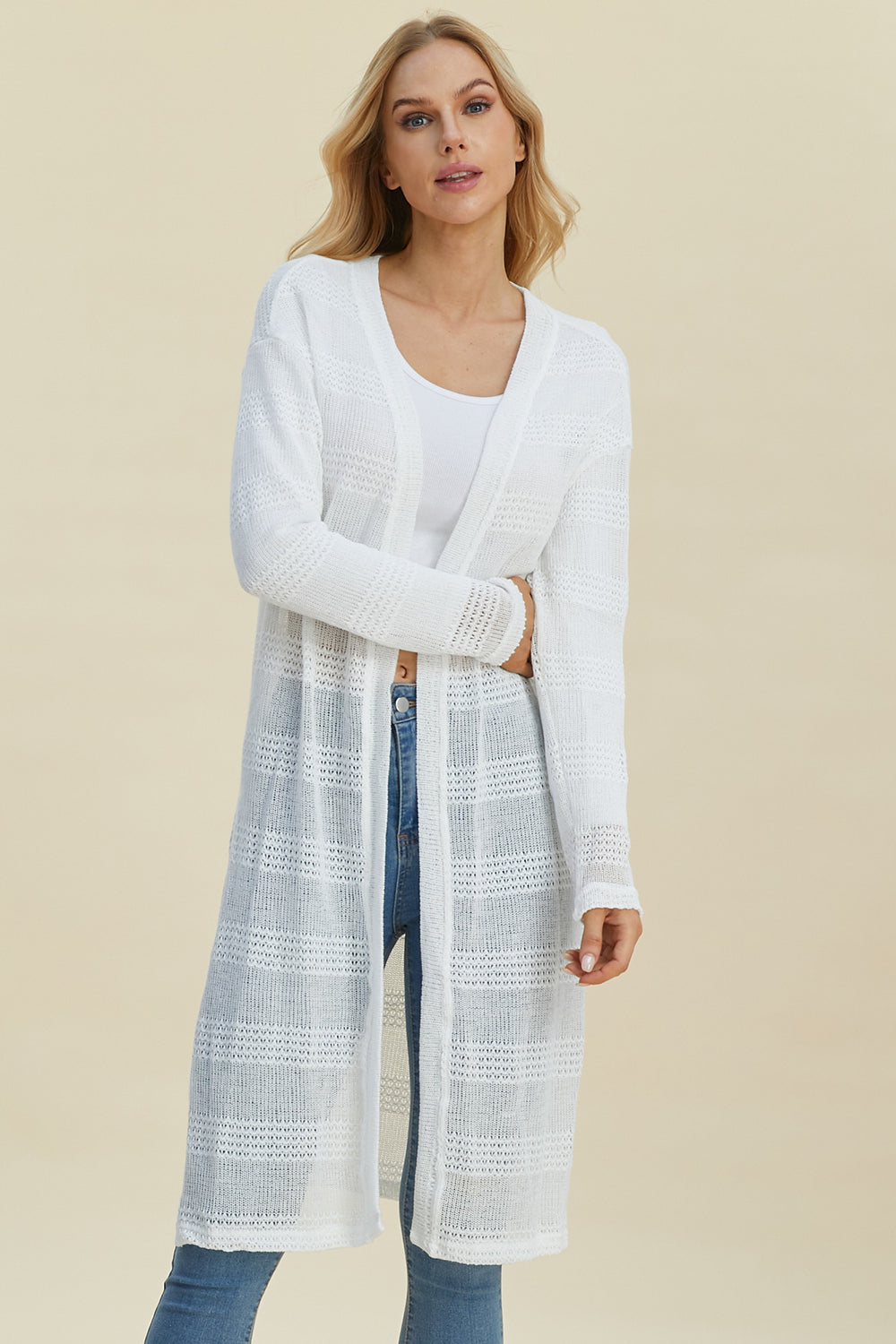 A person wearing the Double Take Full Size Open Front Longline Cardigan in white, featuring an openwork pattern, over a white top and stretch blue jeans, stands against a beige background. They have long, light-colored hair and are looking forward with a neutral expression.