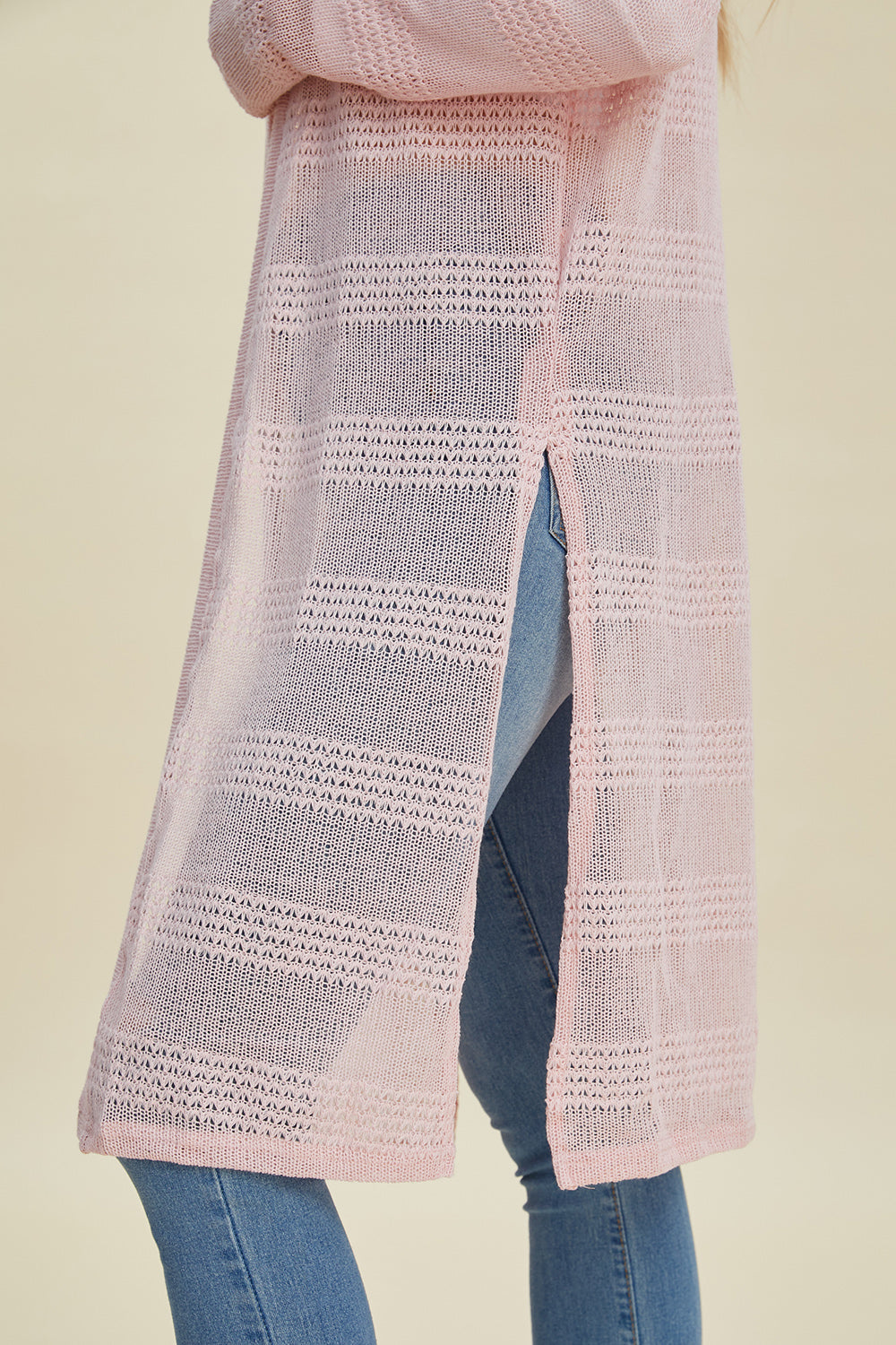 A person wearing blue jeans and the Double Take Full Size Open Front Longline Cardigan, featuring a long, light pink openwork knit design with a vertical slit on the back. The background is a neutral beige color.