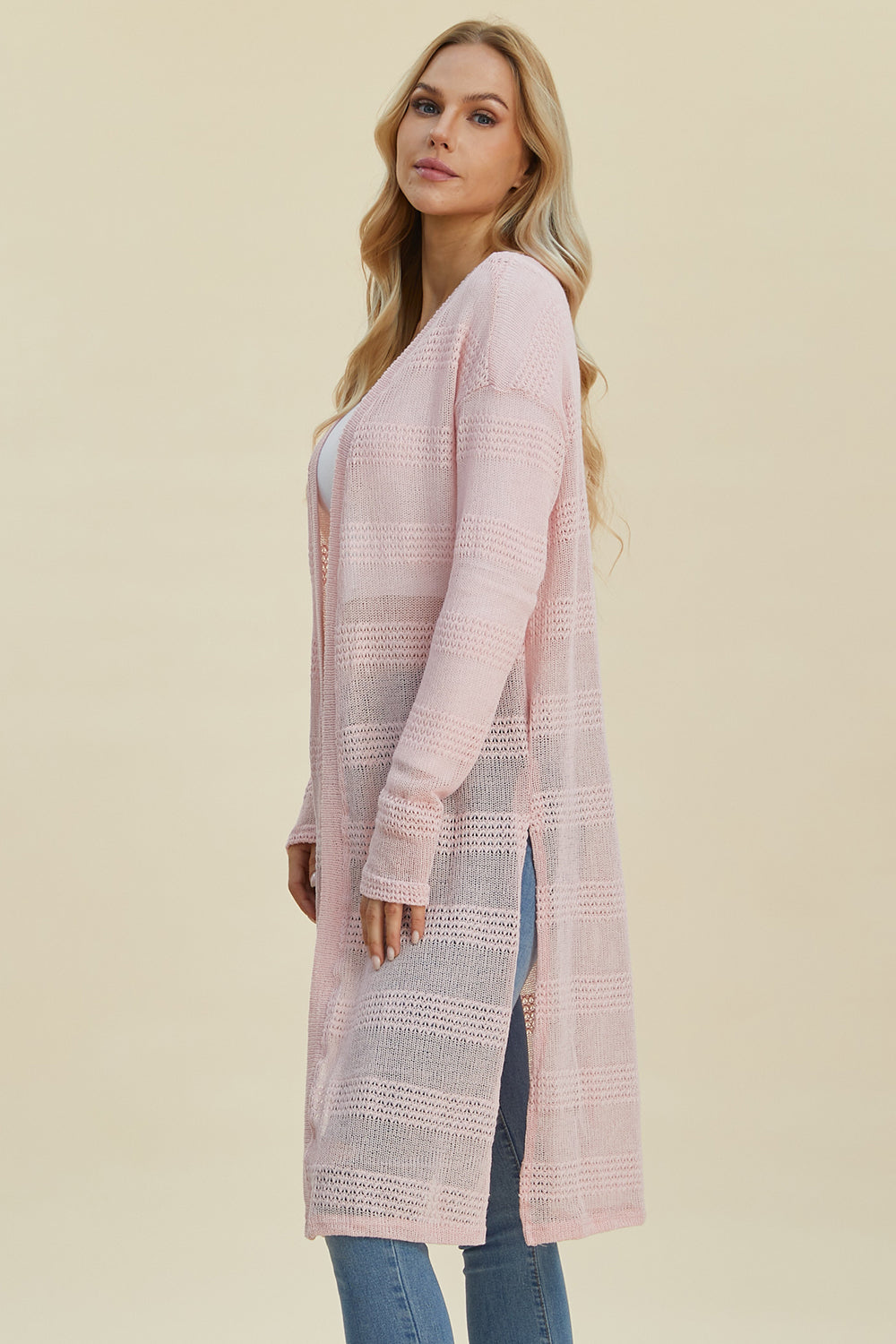 A woman with long blonde hair is wearing the Double Take Full Size Open Front Longline Cardigan, a light pink openwork piece made from a soft polyester blend, draped over a white top and blue jeans as she stands against a neutral background.