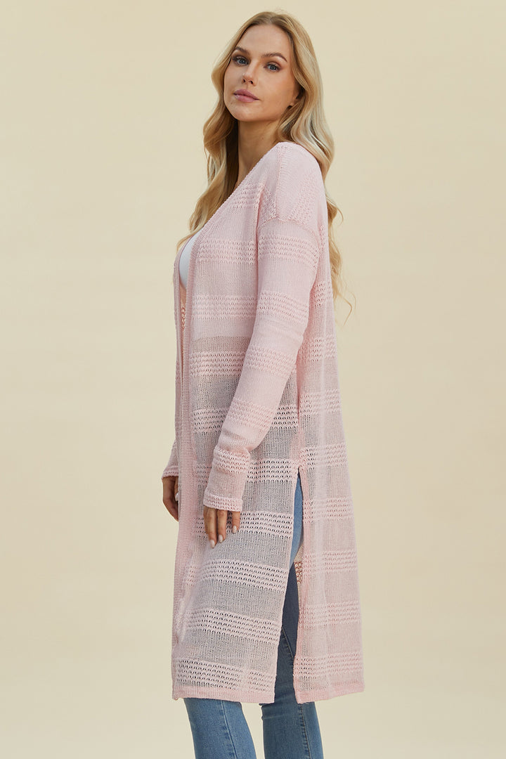 A woman with long blonde hair is wearing the Double Take Full Size Open Front Longline Cardigan, a light pink openwork piece made from a soft polyester blend, draped over a white top and blue jeans as she stands against a neutral background.