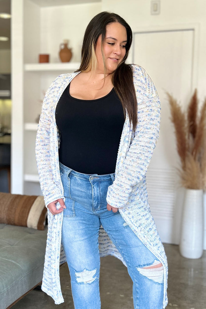 A person with long hair stands indoors wearing a black top and blue ripped jeans, complemented by the Double Take Full Size Open Front Longline Cardigan. A cozy living space with decorative elements is visible in the background.