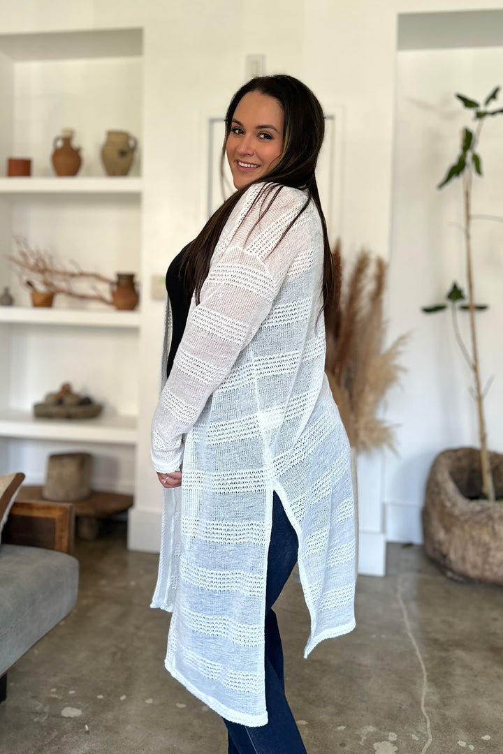 A smiling woman stands in a modern living space, wearing the Double Take Full Size Open Front Longline Cardigan with openwork detailing over a black top and jeans. With her long dark hair cascading down her back, she glances over her shoulder. The room is adorned with minimalist decor, featuring shelves filled with pottery and plants.