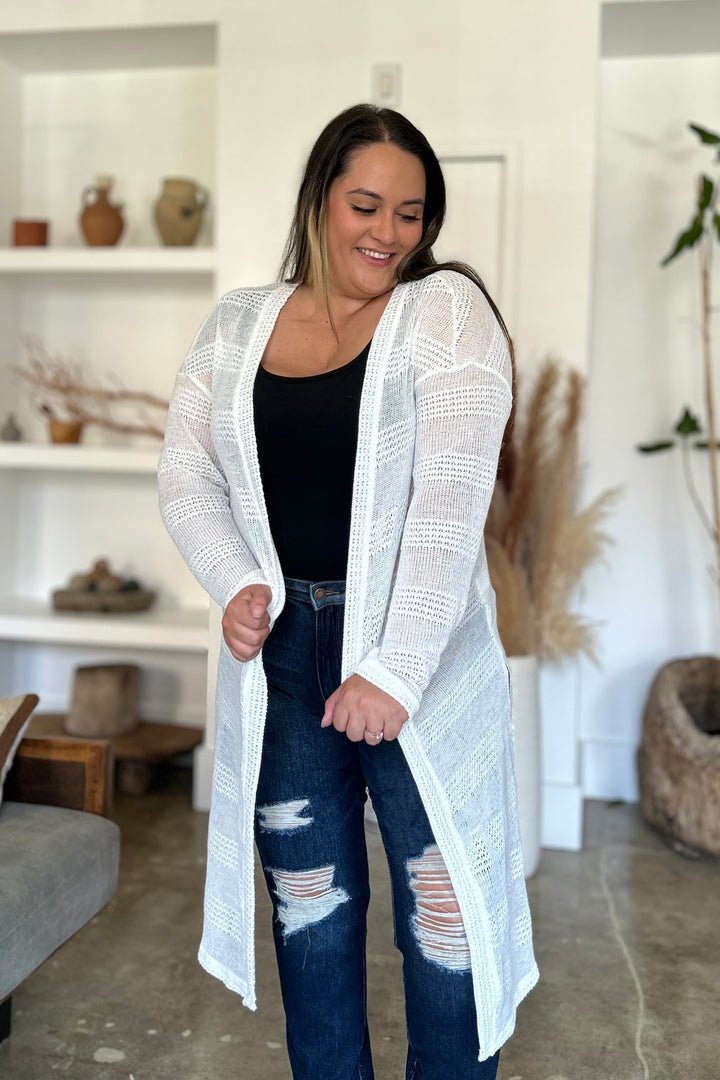 A woman stands in a stylish room, wearing the Double Take Full Size Open Front Longline Cardigan over a black top and ripped jeans. She smiles and looks down, with shelves in the background displaying pottery and decor. Stretching vines add greenery to the corner, enhancing the cozy atmosphere.