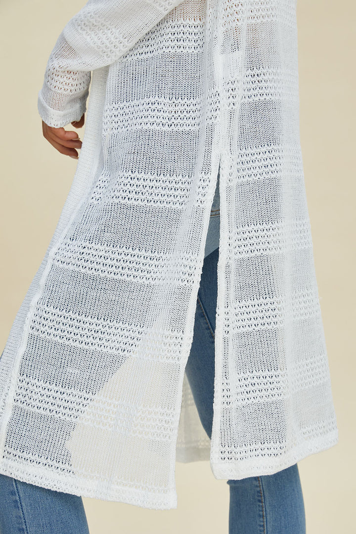 A person is wearing the Double Take Full Size Open Front Longline Cardigan, which is long, white, and features openwork patterns. The polyester cardigan showcases vertical stripes of loose knit designs and gracefully extends past the knees with an open front, paired with blue jeans against a soft beige background.