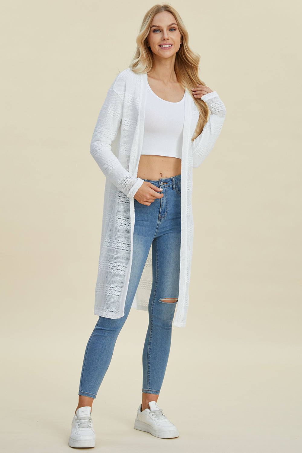 A woman with long blonde hair wears the Double Take Full Size Open Front Longline Cardigan in white, paired with a white crop top and ripped blue jeans. She stands confidently with a slight smile and one hand on her hip against a plain backdrop, completing her look with stretchy white sneakers.