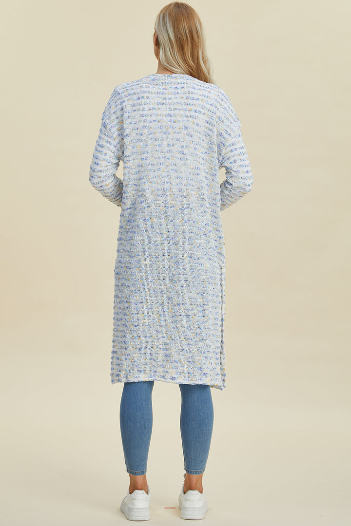 A person with long blonde hair stands with their back to the camera, wearing the Double Take Full Size Open Front Longline Cardigan in light blue and white speckled openwork stretch polyester, paired with blue jeans and white sneakers. The background is a soft beige.