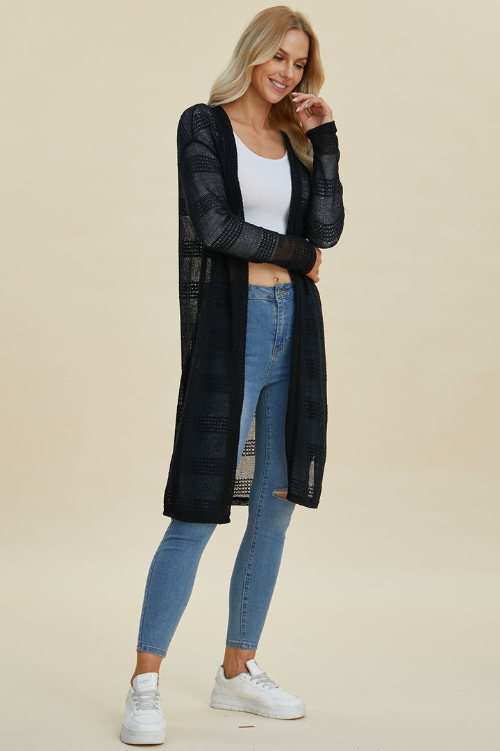 A person is dressed in the Double Take Full Size Open Front Longline Cardigan, a long black openwork piece made from stretch polyester, layered over a white top and blue jeans. They are posing against a plain backdrop while wearing white sneakers.