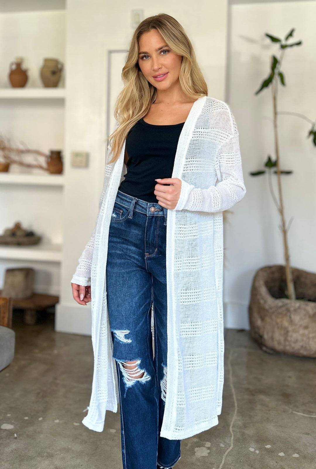 A woman with long blonde hair wearing a black top, distressed jeans, and the Double Take Full Size Open Front Longline Cardigan stands in a modern room with shelves and potted plants.