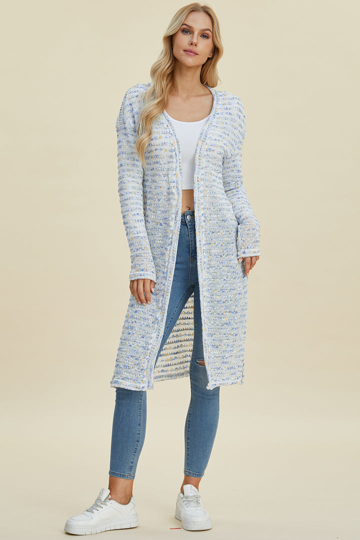 A person with long blonde hair wears the Double Take Full Size Open Front Longline Cardigan, which is a multicolored openwork knit design, over a stretch polyester white tank top. They pair the look with blue jeans and white sneakers while standing on a light background.