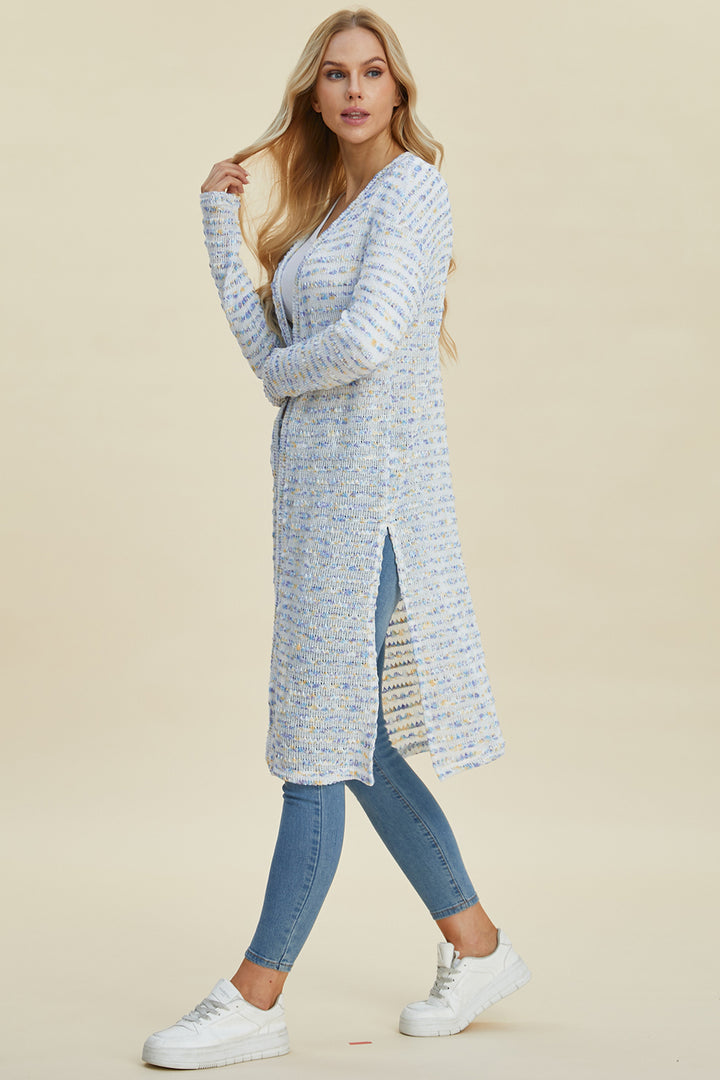 Wearing a Double Take Full Size Open Front Longline Cardigan with a ribbed texture, a person with long blonde hair pairs it with skinny jeans and white sneakers. They are standing against a plain beige background, looking to the side while reaching one hand towards their hair.