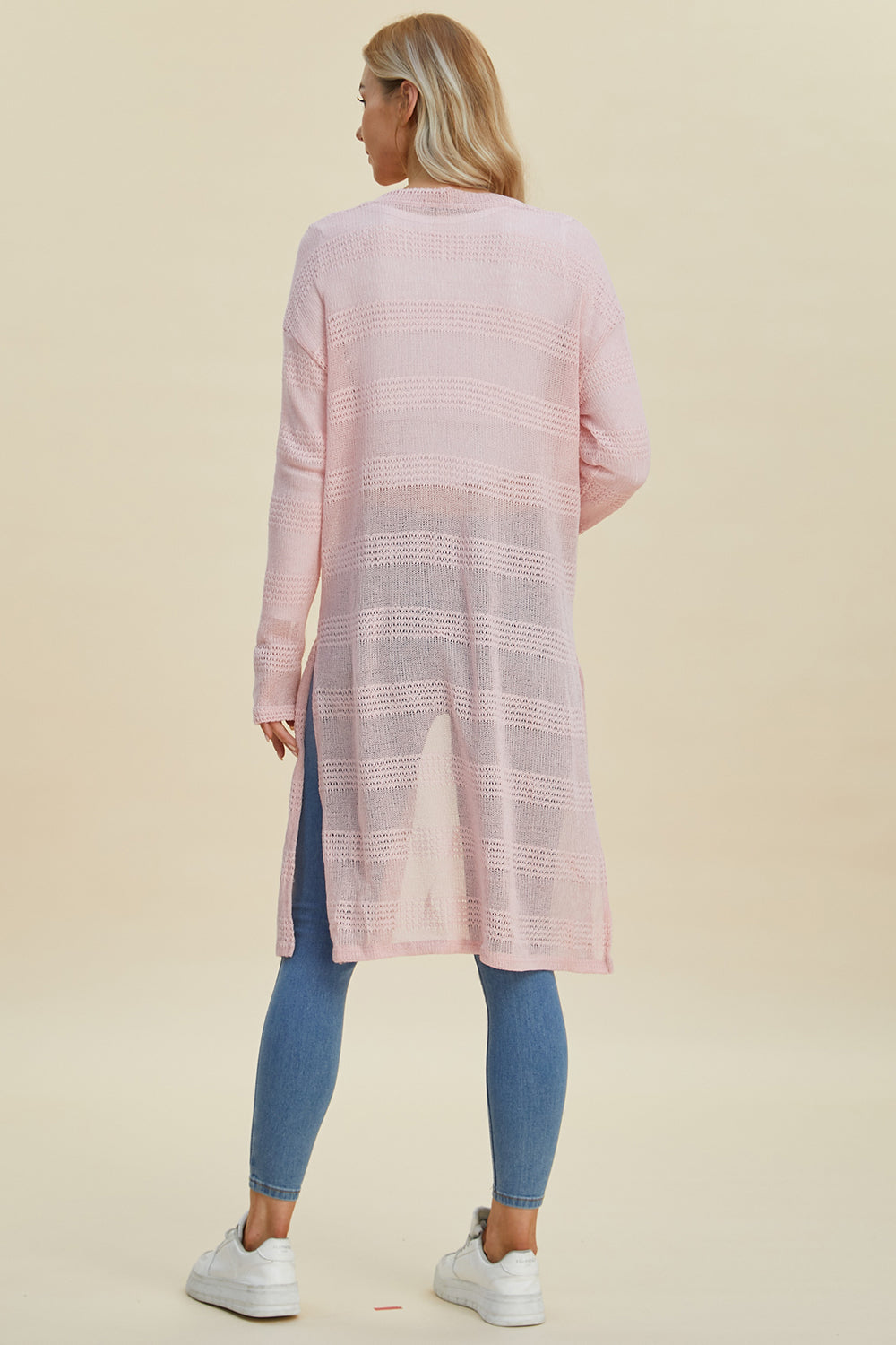 A person with long hair is wearing the Double Take Full Size Open Front Longline Cardigan in light pink, featuring an openwork textured pattern. They're pairing it with blue jeans and white sneakers. The cardigan has side slits and offers a hint of stretch as they stand facing away against a neutral background.