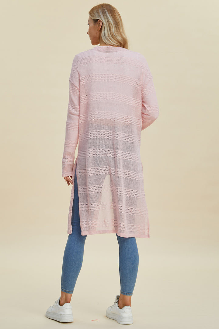 A person with long hair is wearing the Double Take Full Size Open Front Longline Cardigan in light pink, featuring an openwork textured pattern. They're pairing it with blue jeans and white sneakers. The cardigan has side slits and offers a hint of stretch as they stand facing away against a neutral background.