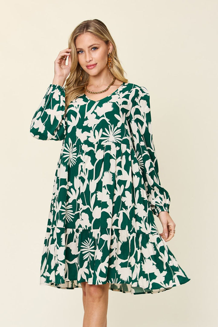 A woman wearing the Double Take Full Size Printed Ruffle Hem Dress with Pocket, featuring a green and white floral design on rayon fabric and long sleeves, stands against a plain background. She touches her hair while accessorizing with minimal jewelry. The dress showcases a tiered skirt design and subtle ruffled details that enhance its elegant appearance.