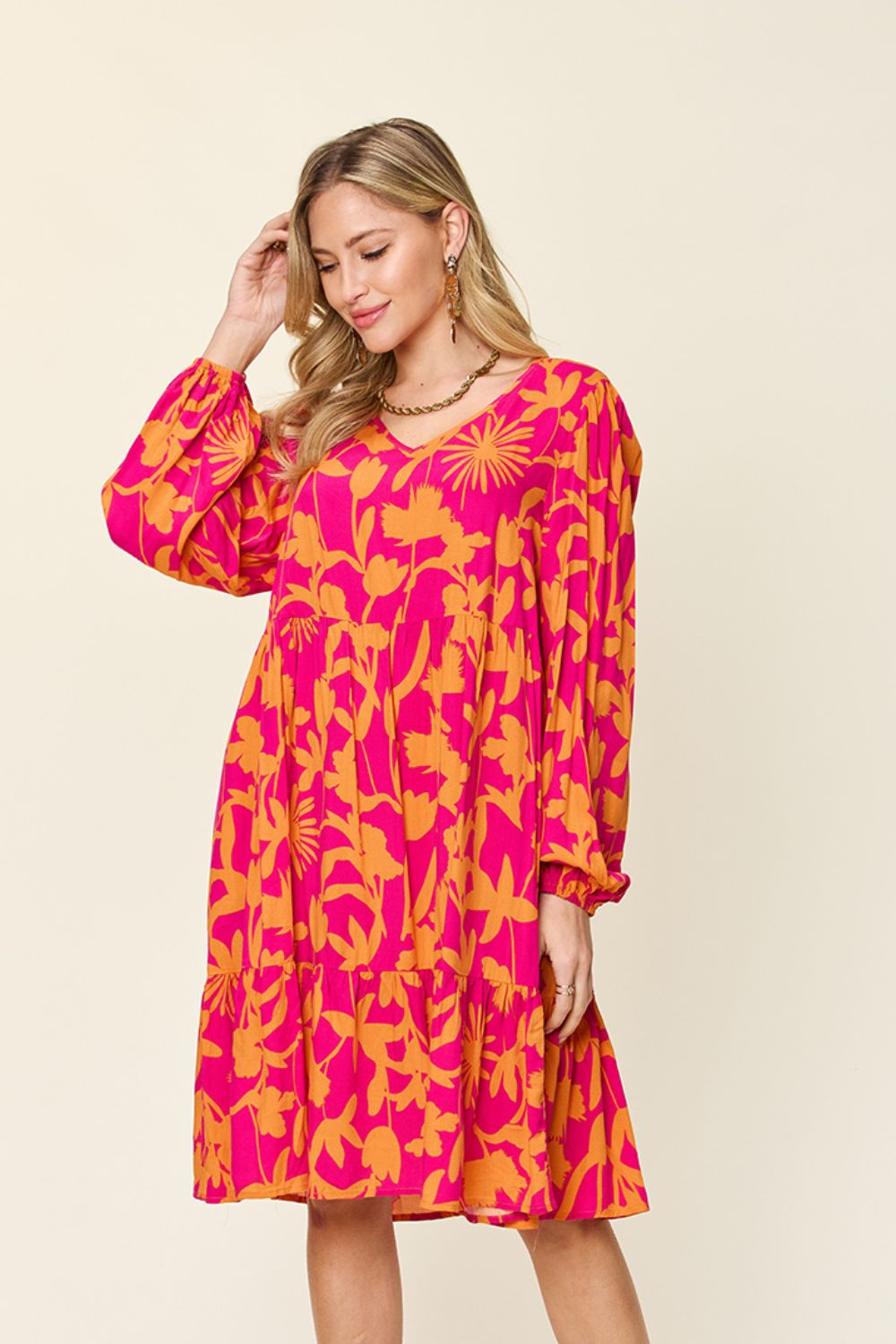 A woman in the Double Take Full Size Printed Ruffle Hem Dress with Pocket, featuring a pink base adorned with yellow floral patterns, poses against a light background. Crafted from soft rayon, the dress highlights long, loose sleeves and a flowing ruffled design. She complements her look with earrings and gently touches her hair, smiling softly.