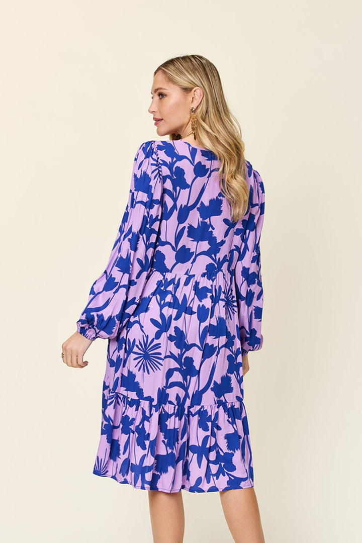 A woman with long blonde hair stands with her back turned, wearing the Double Take Full Size Printed Ruffle Hem Dress with Pocket. The dress is decorated with a purple and blue floral pattern and made of rayon, featuring long sleeves and subtle ruffled details. She looks to her right against a plain, light-colored wall backdrop.