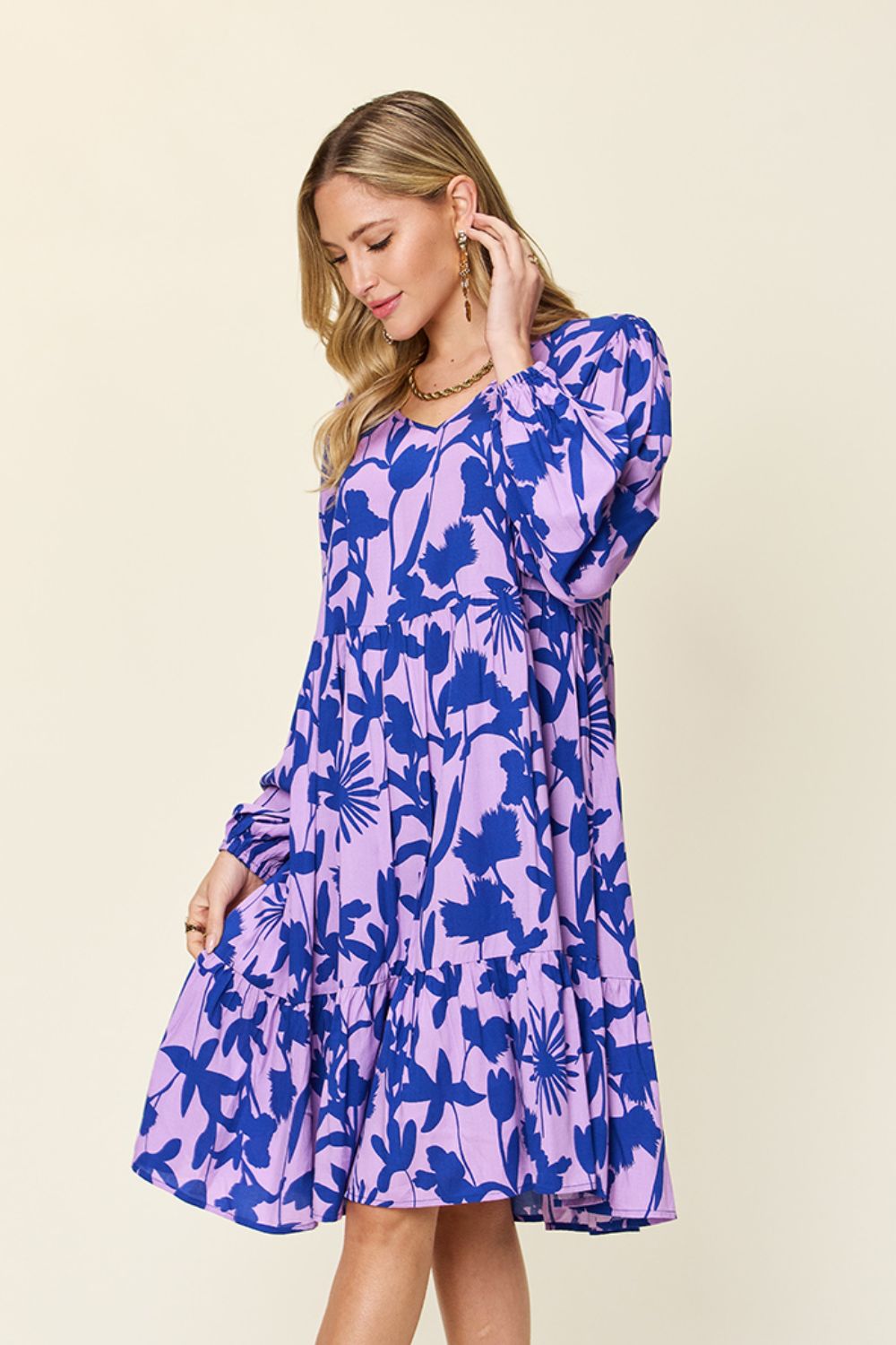 A woman wearing the Double Take Full Size Printed Ruffle Hem Dress with a pocket in purple, featuring blue floral patterns, stands in profile looking down. She has long blonde hair and gently holds one side of the dress with her hand. The background is a plain light color.