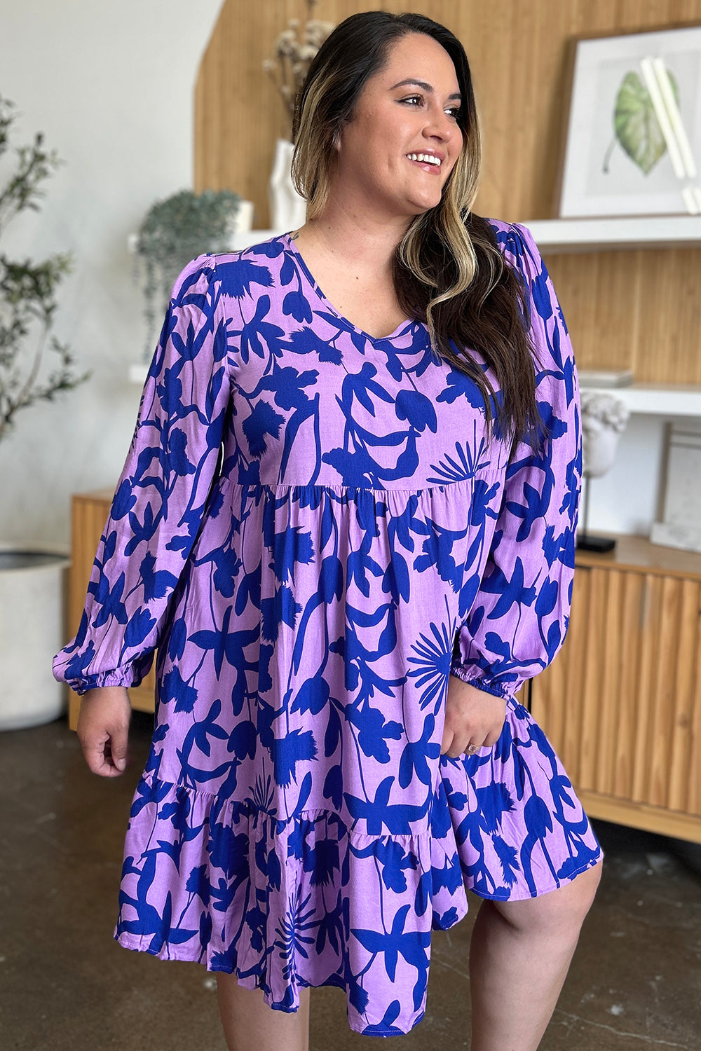 A person is smiling while wearing the Double Take Full Size Printed Ruffle Hem Dress with Pocket, featuring a purple and blue floral pattern. They are standing indoors, surrounded by wooden shelves and green plants in the background.