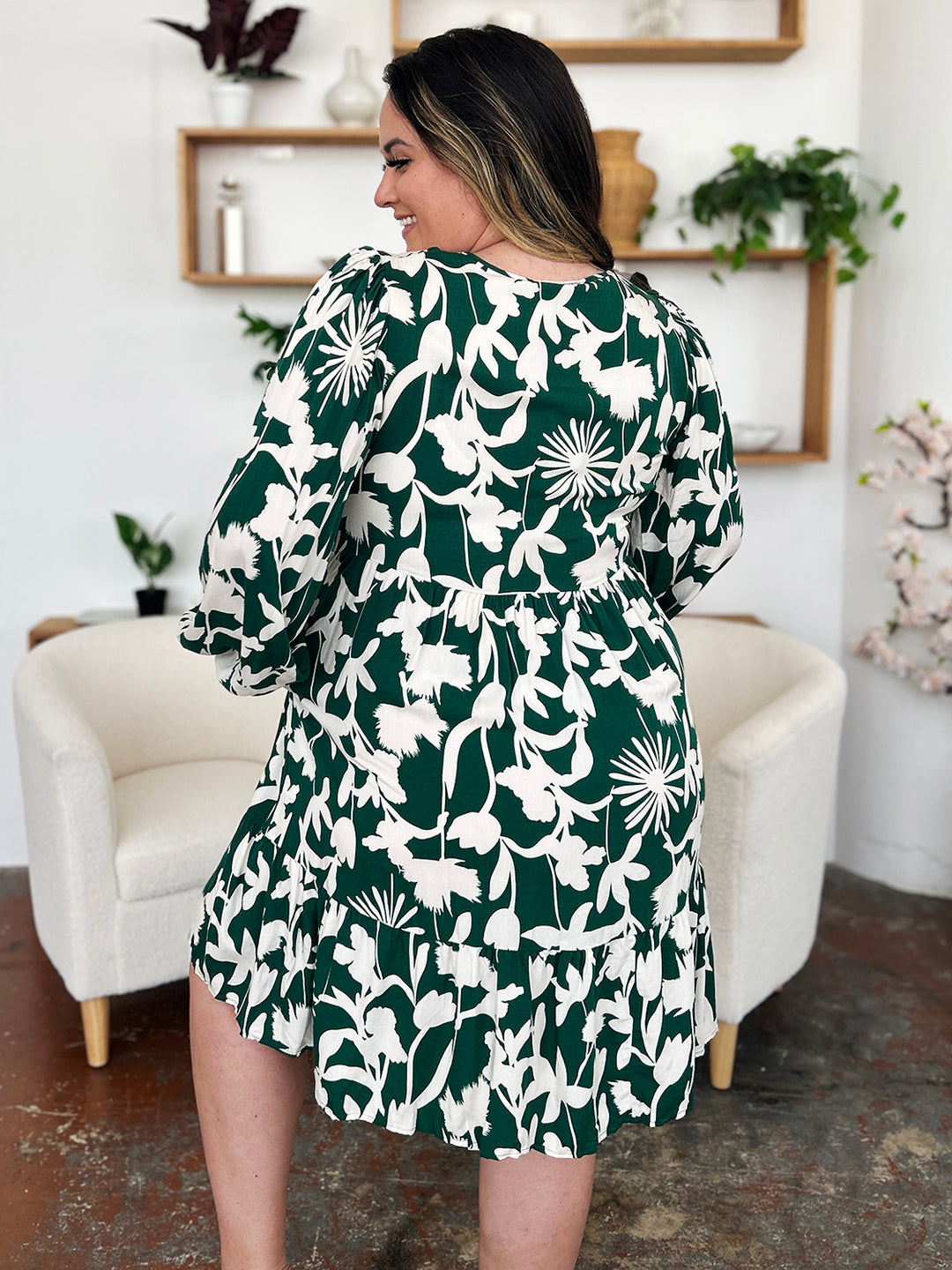 A woman with long hair stands confidently in a room, wearing the Double Take Full Size Printed Ruffle Hem Dress with Pocket in green floral. Her back is turned, highlighting the ruffled pattern. The room features white chairs, plants on shelves, and decorative items on the walls.