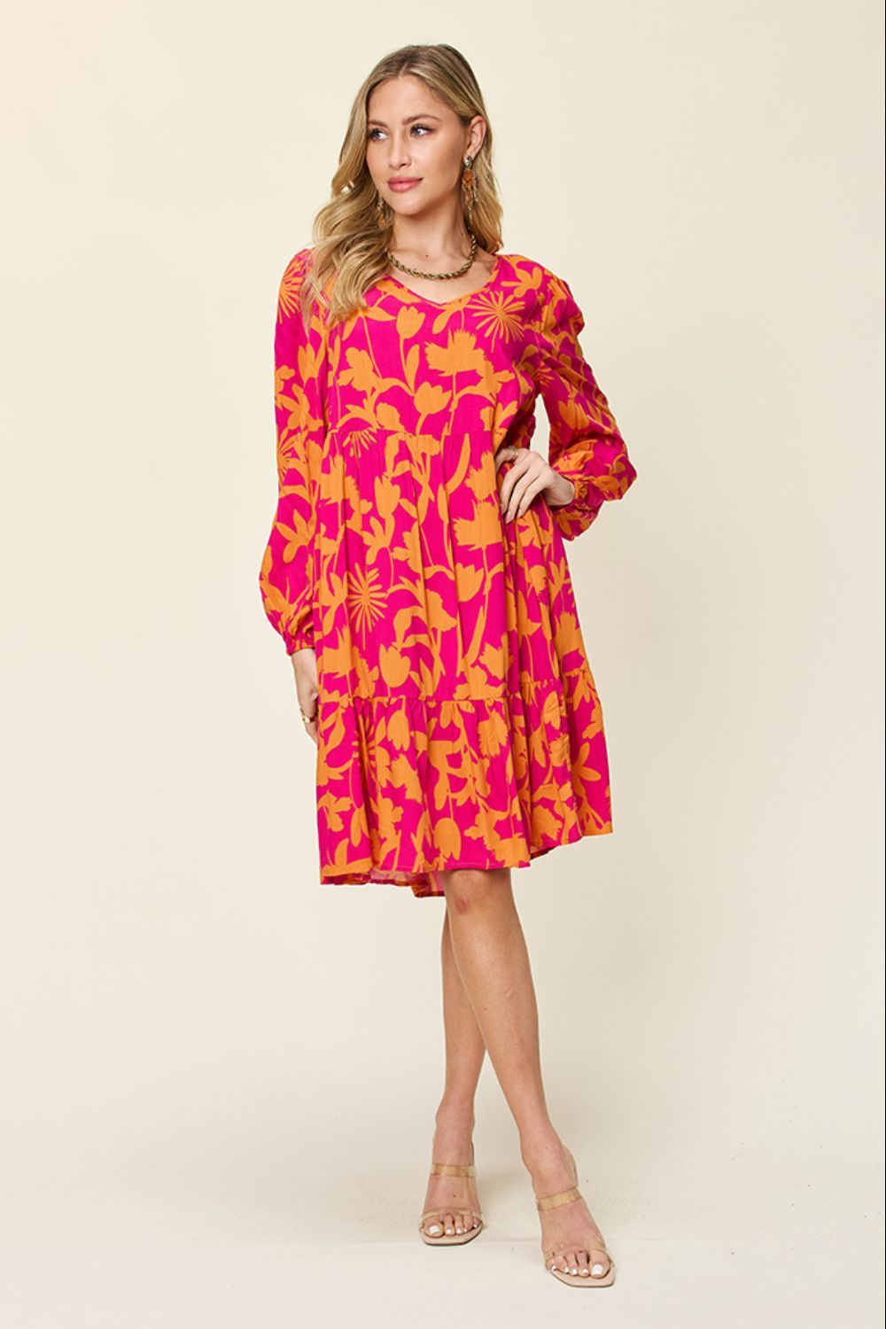 A woman poses in the Double Take Full Size Printed Ruffle Hem Dress with Pocket, a pink rayon dress adorned with yellow floral patterns. The dress features long sleeves and a ruffled, flowing skirt. She is wearing beige heels and has long blonde hair against a plain off-white background.