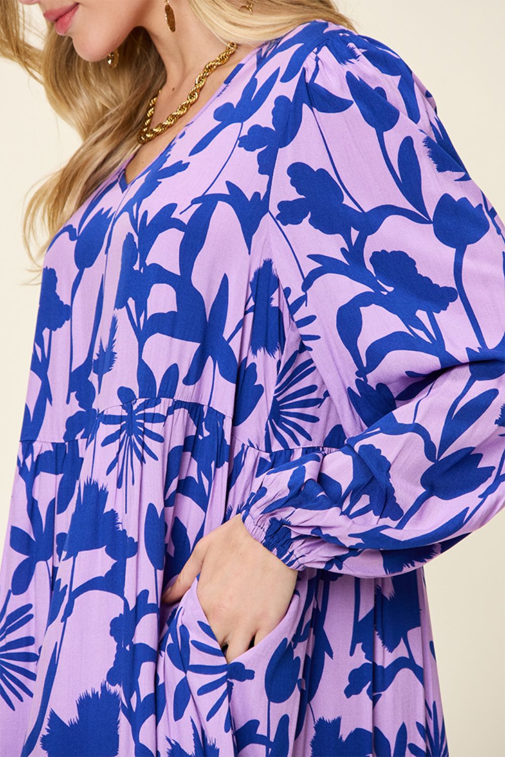 The individual is dressed in the Double Take Full Size Printed Ruffle Hem Dress with Pocket, showcasing bold blue floral patterns on a purple background. Made from soft rayon, this dress features long sleeves and a V-neckline, offers a relaxed fit, and includes convenient side pockets where the wearer casually rests their hand.