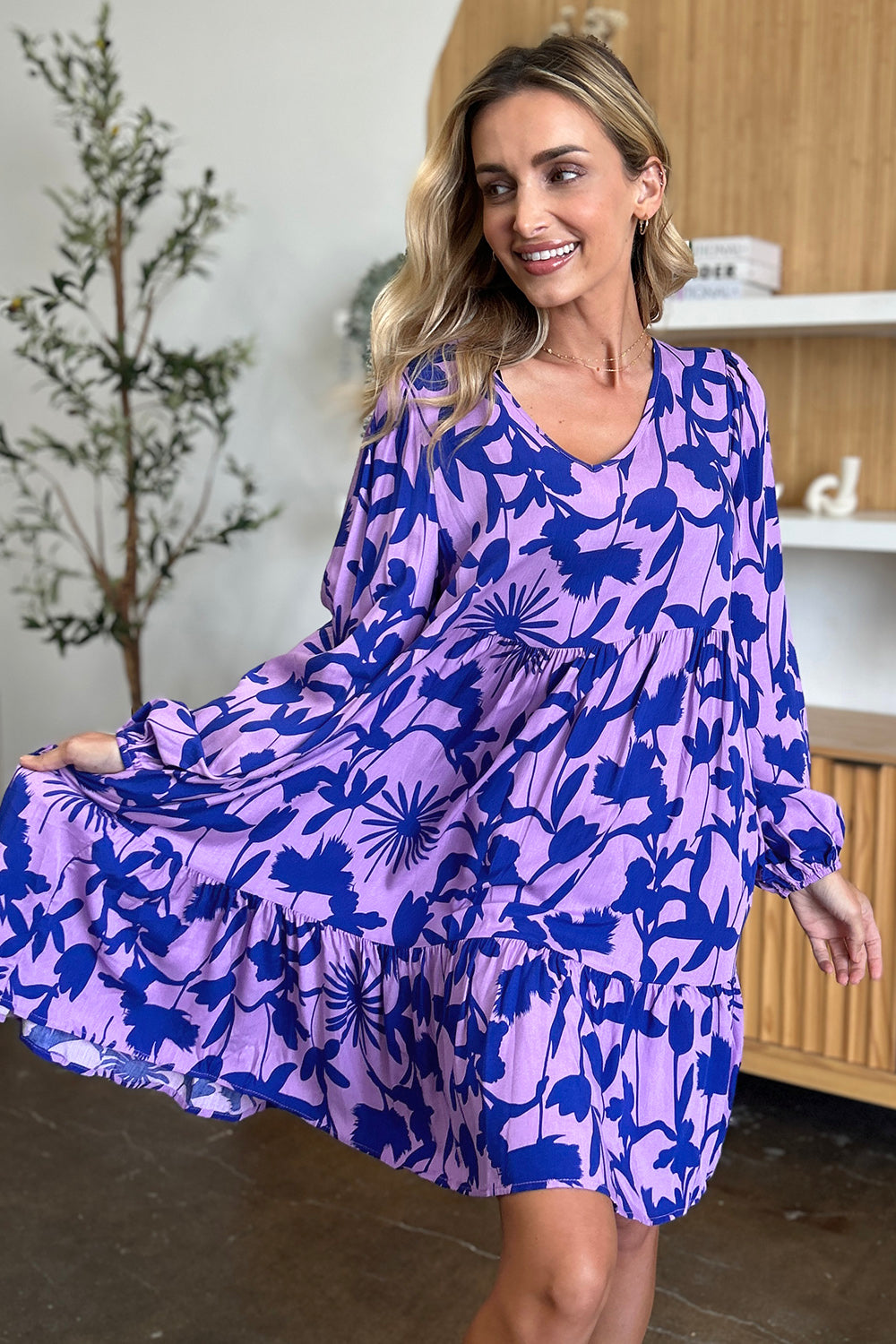 A woman smiles while wearing the "Double Take Full Size Printed Ruffle Hem Dress with Pocket," a rayon dress featuring a ruffled hem and a blue floral pattern on purple fabric. She poses indoors near a small tree and wooden shelves, gently holding the side of her dress with her left hand.