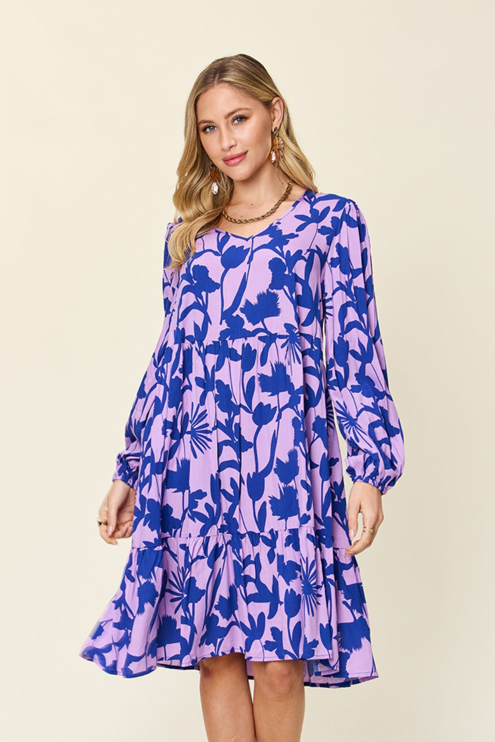 A woman wears the Double Take Full Size Printed Ruffle Hem Dress with Pocket, a stunning purple rayon piece adorned with a blue floral pattern. The dress features long sleeves and a ruffled hem, effortlessly combining style and convenience with its handy pockets. Her long, wavy hair beautifully complements her hoop earrings and necklace against a plain, light background.