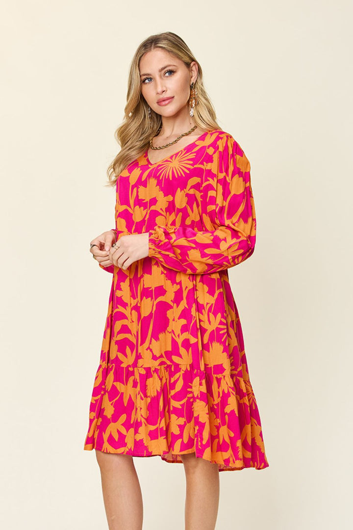 A woman wearing the Double Take Full Size Printed Ruffle Hem Dress with Pocket in vibrant pink and yellow floral stands against a beige background. The ruffled hem adds an elegant touch as she poses with one hand lightly holding the other, her long hair cascading to one side. The dress's pocket details subtly enhance her ensemble, which is complemented by dangling earrings.