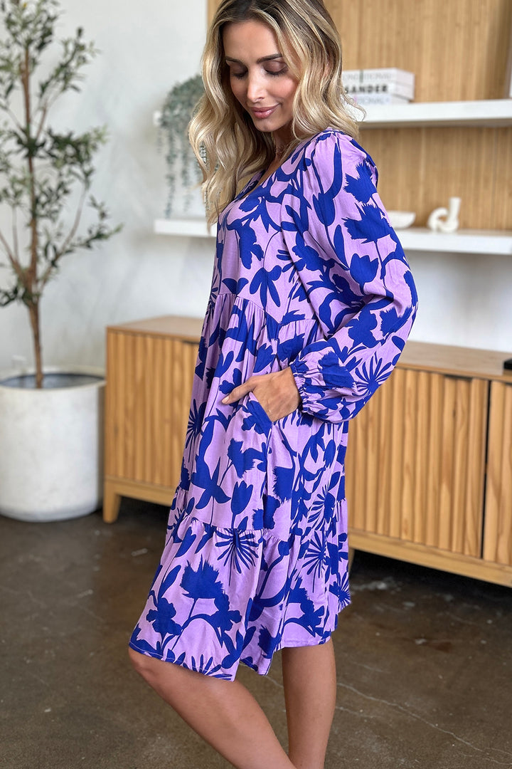 A woman wearing the Double Take Full Size Printed Ruffle Hem Dress with Pocket, featuring a vibrant blue floral pattern on purple rayon, stands gracefully in a room. Her hands are tucked into the dress's ruffled pockets as she gazes down with a gentle smile, enveloped by the warm tones of a wooden cabinet and the freshness of a potted plant.