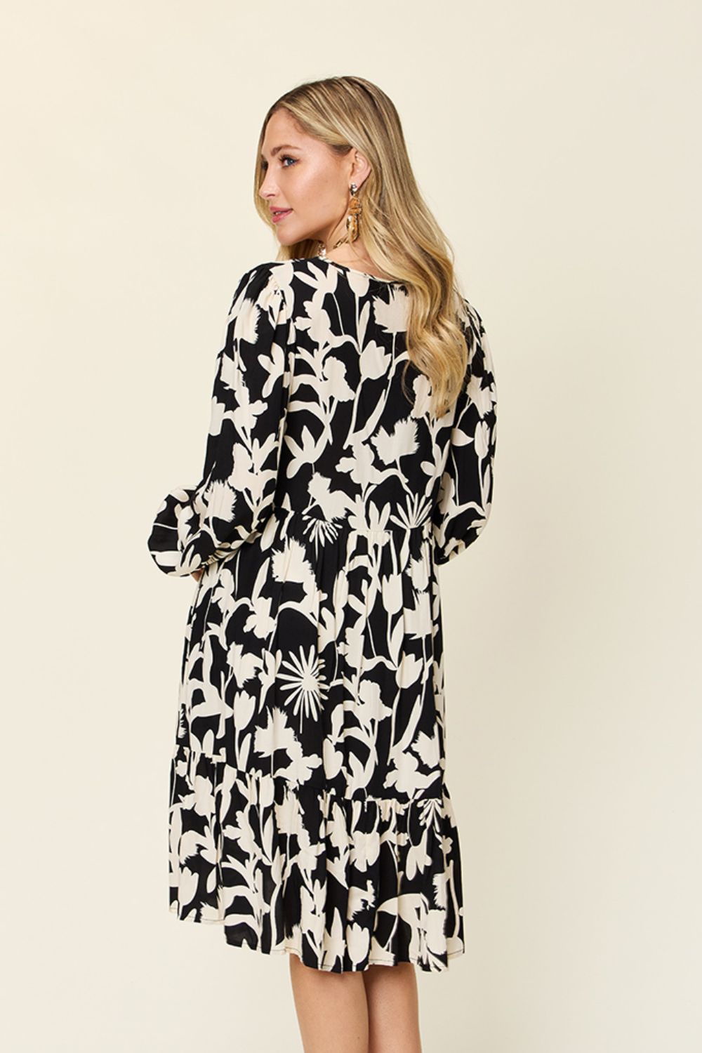 A woman with long blonde hair, wearing gold earrings and the Double Take Full Size Printed Ruffle Hem Dress with Pocket—an elegant black dress adorned with a white floral pattern—stands facing away. The ruffled hem sways as she glances over her shoulder against a light background.