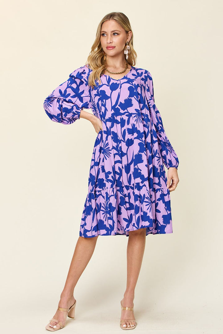 A woman models the Double Take Full Size Printed Ruffle Hem Dress with Pocket against a light background. The dress, adorned with a blue and purple floral print, is crafted from soft rayon and features ruffled detailing, long sleeves, and a knee-length hem. She pairs the outfit with dangling earrings and beige high-heeled sandals, while discreet pockets lend subtle functionality.