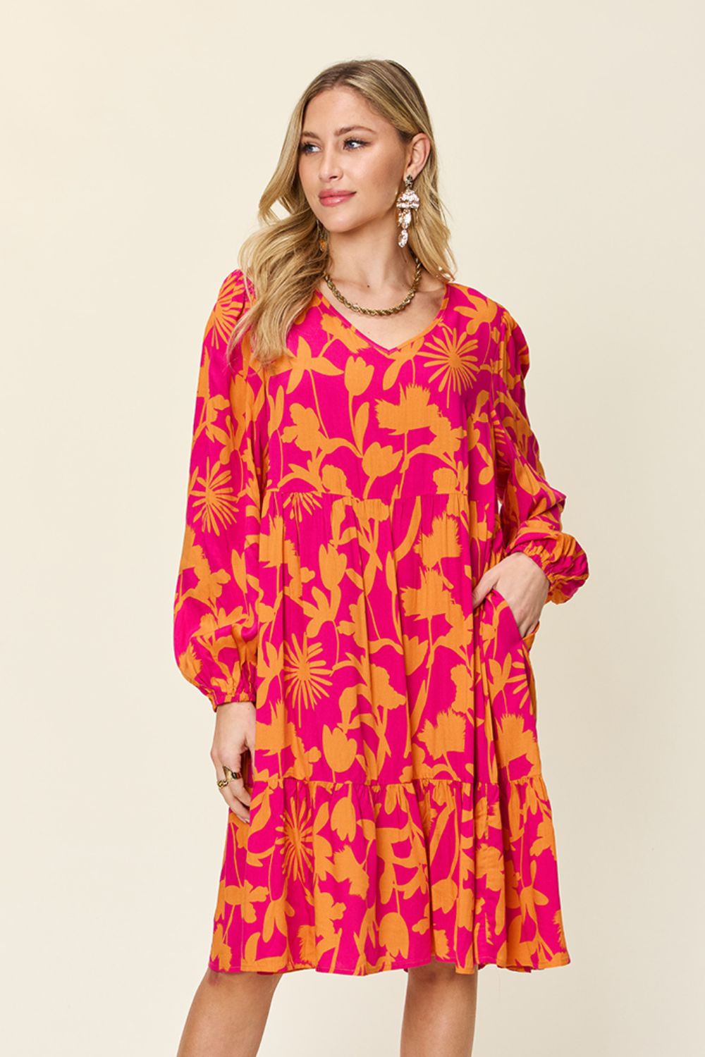 A woman models the "Double Take Full Size Printed Ruffle Hem Dress with Pocket" in a striking pink and orange floral rayon pattern against a simple backdrop. This knee-length dress is designed with long sleeves and features elegant ruffled accents. She accessorizes the ensemble with earrings and a necklace.