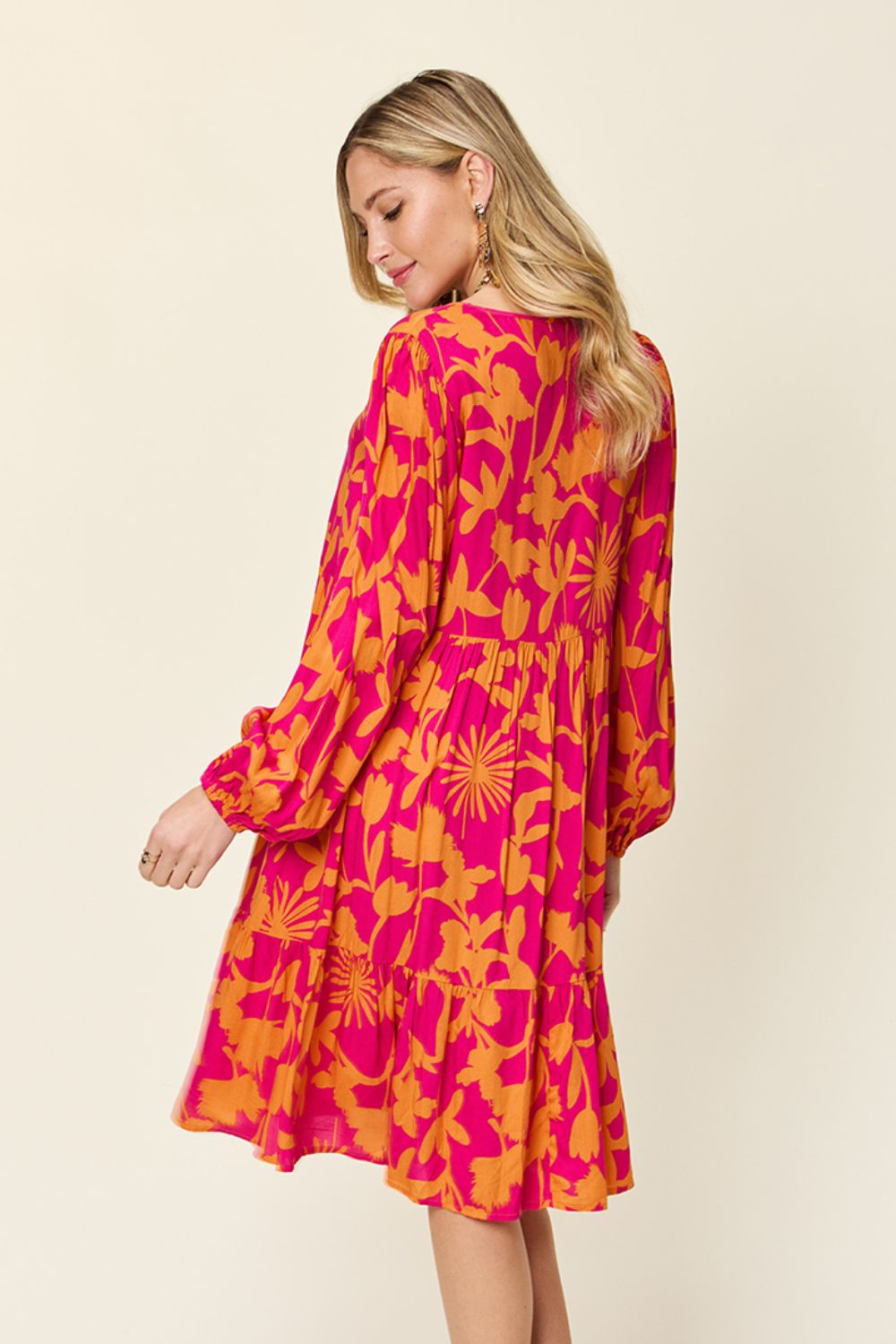 A woman wearing the Double Take Full Size Printed Ruffle Hem Dress with Pocket in a vibrant pink and orange floral pattern is standing, looking down over her shoulder. The long-sleeved design features subtle ruffled details and practical pockets, set against a light and neutral background.