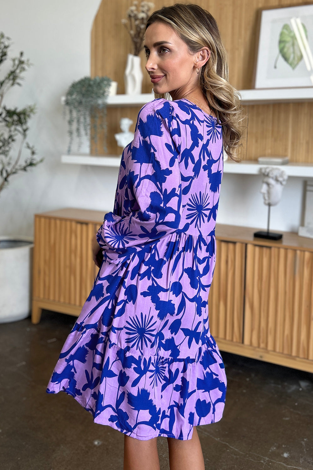 A woman in the Double Take Full Size Printed Ruffle Hem Dress with pockets stands in a room filled with wooden furniture and decorative plants. Her dress, crafted from soft rayon, features a purple hue adorned with blue floral patterns. She is turned slightly, looking over her shoulder.