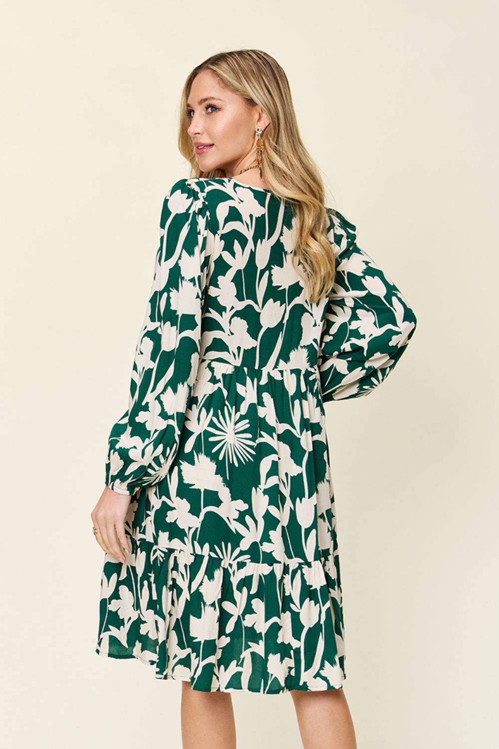 A woman with long blonde hair is seen standing sideways, wearing the Double Take Full Size Printed Ruffle Hem Dress with Pocket. The dress is crafted from rayon and showcases a pocketed design with white floral patterns on a green background, featuring long sleeves and a gracefully flowing skirt. She stands against a plain, light backdrop.