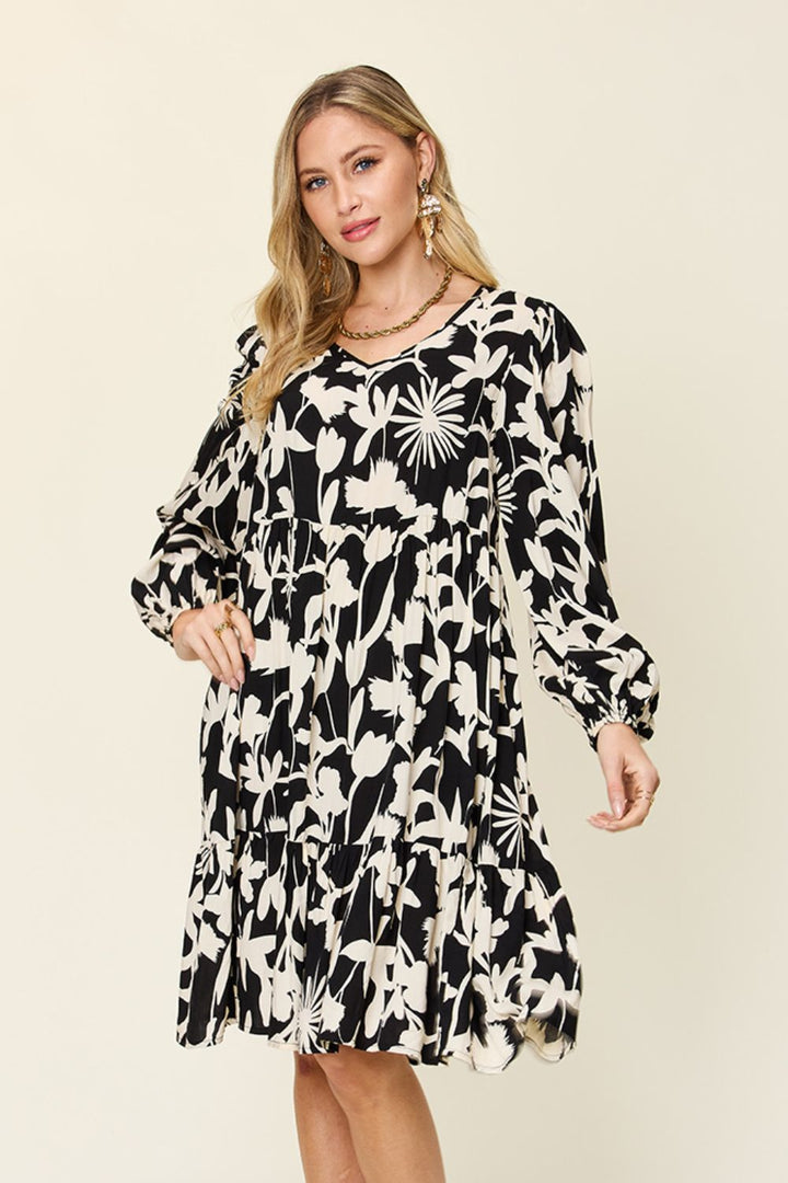 A woman with long hair is wearing the Double Take Full Size Printed Ruffle Hem Dress with Pocket, designed in a striking black and white floral pattern and crafted from soft rayon. The dress features long sleeves and subtle ruffled details. She smiles warmly, posing against a plain background.