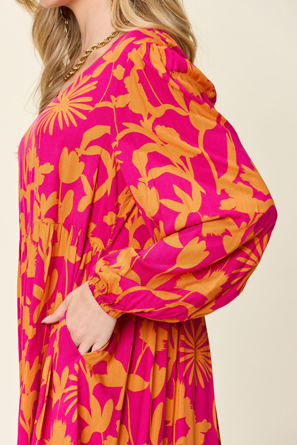 A person is wearing the Double Take Full Size Printed Ruffle Hem Dress with Pocket, which showcases a vibrant pink color with bold orange floral patterns. This rayon dress features long sleeves, a relaxed fit, and ruffled accents, emphasizing its colorful design.