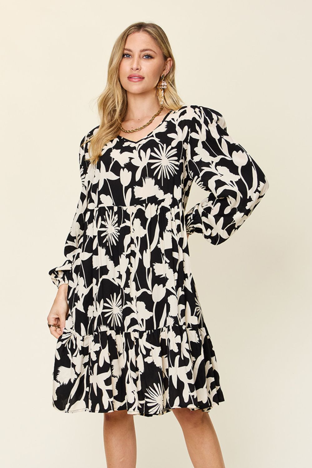 A woman is elegantly wearing the Double Take Full Size Printed Ruffle Hem Dress with a stylish black and white floral design, featuring convenient pockets and long sleeves. The rayon dress boasts a tiered ruffle hem, beautifully accessorized by gold earrings and a necklace. She poses gracefully against a plain beige background.