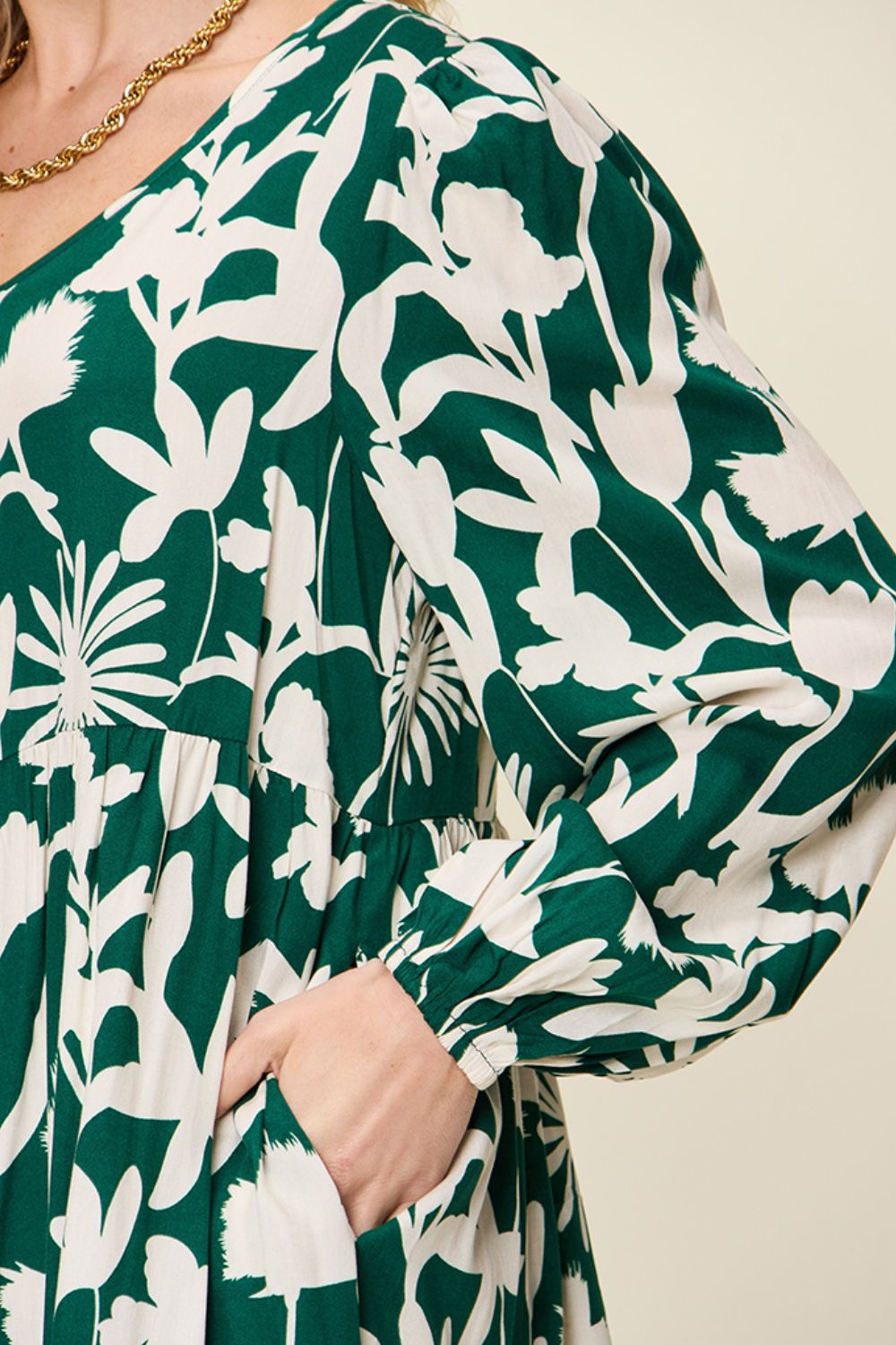 A close-up of an individual wearing the Double Take Full Size Printed Ruffle Hem Dress with Pocket, showcasing its green rayon fabric adorned with a white floral pattern. The pocketed dress features long, slightly puffed sleeves, allowing the person to casually slip a hand into one pocket, while a gold chain necklace can be partially seen.