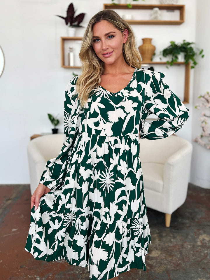 A woman stands in a room wearing the Double Take Full Size Printed Ruffle Hem Dress with Pocket. This green dress, adorned with white floral patterns and crafted from soft rayon, features a stylish ruffled hem. She poses with one hand holding the side of her dress, while a beige armchair and shelves filled with plants and decorative items create an inviting background.