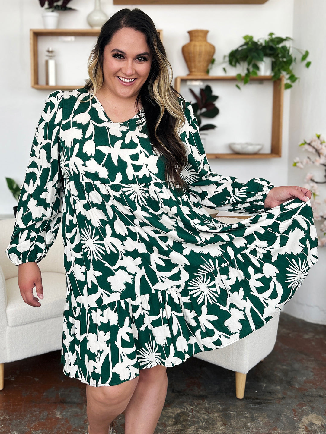 Wearing the Double Take Full Size Printed Ruffle Hem Dress with Pocket, a person smiles as they hold the hem, showing off its subtle pocketed design. They pose indoors with white shelves, lush plants, and a cream-colored chair in the background.