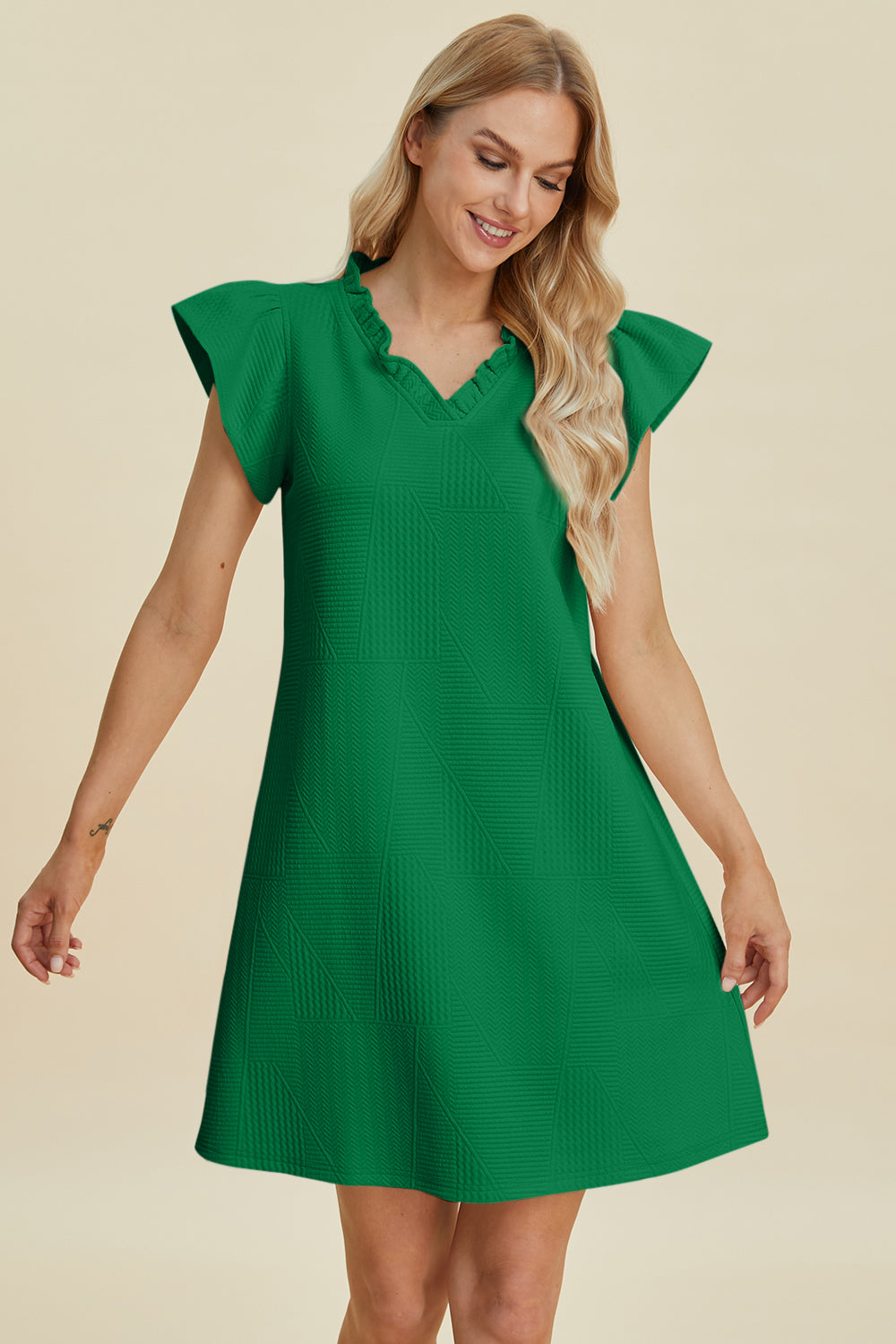 A smiling woman stands against a neutral background in a green Double Take Full Size Ruffled V-Neck Cap Sleeve Dress, made of soft polyester. Her long, wavy blonde hair flows effortlessly as she looks relaxed and happy. The dress is easy-care for machine wash cold.