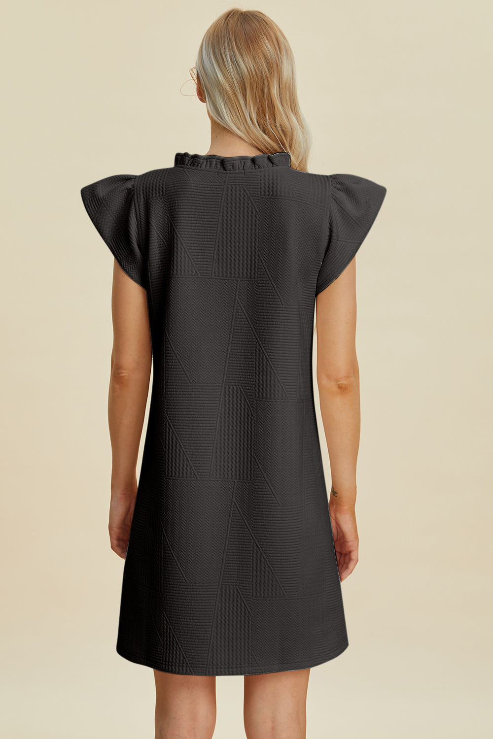 A person with long blonde hair, viewed from the back, wears the Double Take Full Size Ruffled V-Neck Cap Sleeve Dress against a beige background. The black textured dress is polyester with short cap sleeves, offering basic style and elegance that can be easily machine washed cold.