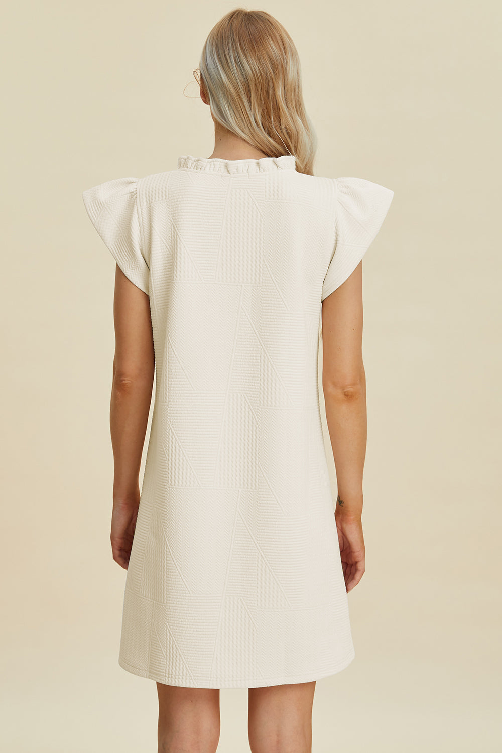 A person with long blonde hair faces away from the camera in a cream Double Take Full Size Ruffled V-Neck Cap Sleeve Dress, notable for its textured polyester fabric and ruffled shoulders. This basic, elegant piece is machine washable for easy care. The backdrop is a simple neutral shade.