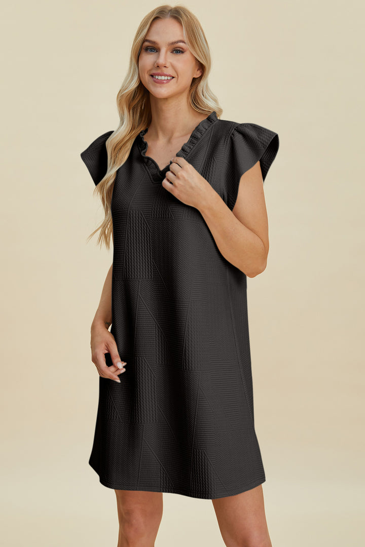 A woman with long blonde hair smiles while wearing a knee-length, black ruffled V-neck cap sleeve dress crafted from polyester for easy care. The dress features a textured pattern and can be machine washed cold. She stands against a neutral background.