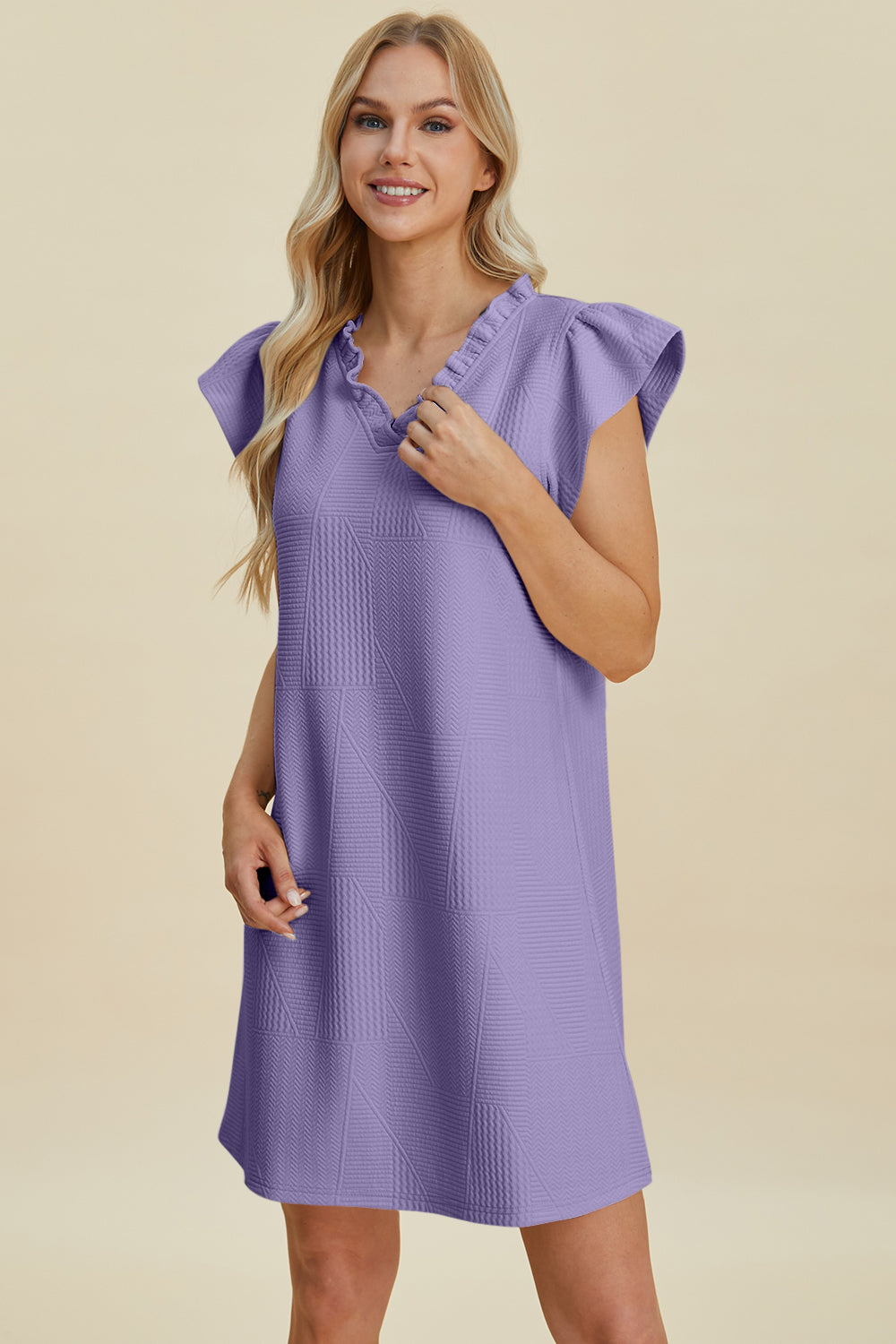 A person with long blonde hair smiles while wearing a Double Take Full Size Ruffled V-Neck Cap Sleeve Dress, made of purple textured polyester with ruffled sleeves, standing out against the plain, soft beige background.