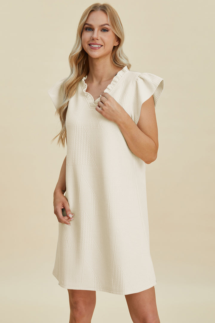 A woman with long blonde hair wears the Double Take Full Size Ruffled V-Neck Cap Sleeve Dress, a short, cream-colored polyester dress with ruffled sleeves and textured pattern. Smiling, she stands against a beige background. The basic-style dress is best cared for by machine washing cold.