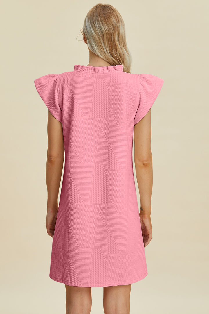 A person with long blonde hair faces away from the camera, wearing a pink Double Take Full Size Ruffled V-Neck Cap Sleeve Dress. Combining style and practicality, its made of polyester, machine washable cold. The background is plain beige.