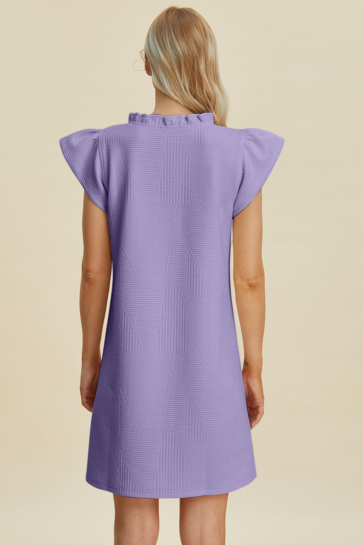 A person with long blonde hair wears the Double Take Full Size Ruffled V-Neck Cap Sleeve Dress, a purple textured piece made from soft polyester. With short flared sleeves, it stands out against a light beige background and is easy to machine wash cold.