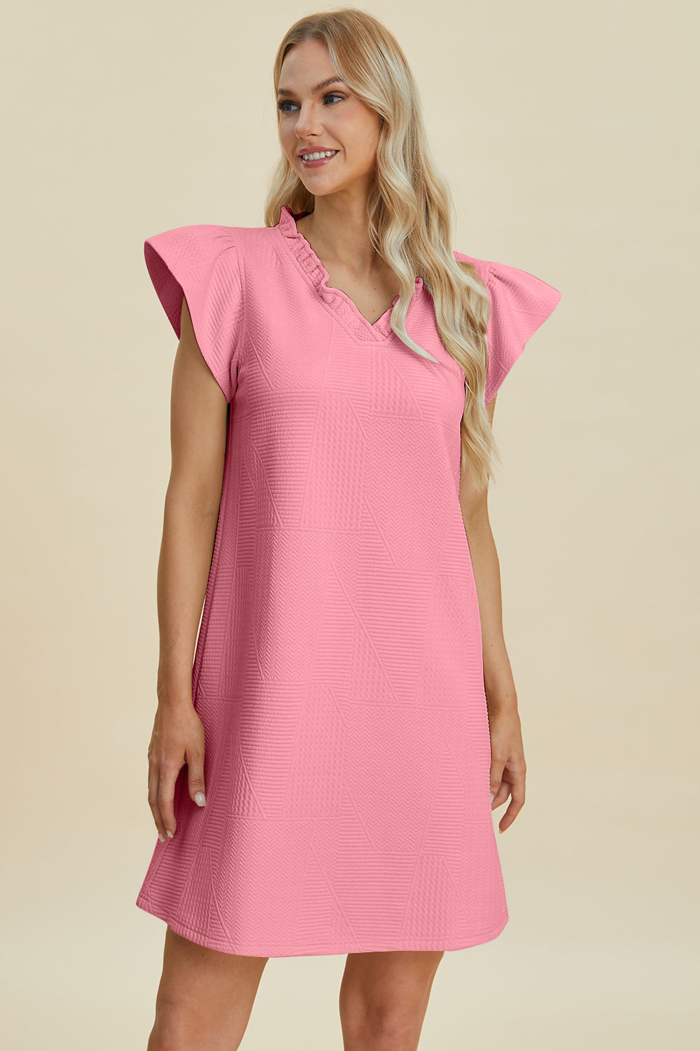 A woman with long blonde hair wears the Double Take Full Size Ruffled V-Neck Cap Sleeve Dress in pink. She poses against a light beige background, smiling with her hands by her side, exuding timeless elegance and chic style.